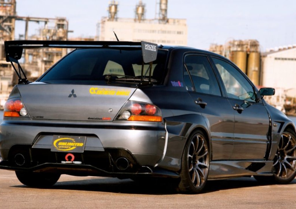 Rear view of a modified grey Mitsubishi Lancer Evolution featuring a widebody kit, Voltex GT wing, and "Unlimited Works" decals, symbolizing the high tuning potential of the 4G63 engine